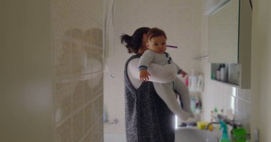 Anne, bebeğini banyoda taşırken dişlerini fırçalarken birden fazla iş yapıyor. Bebek, lavabodaki nesneleri merakla inceleyerek öne doğru eğilir.