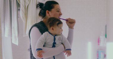 Anne banyoda birden fazla iş yapıyor, bebeğini tutarken dişlerini fırçalıyor. Bebek, sabah rutininden büyülenmiş bir şekilde çevreye merakla bakıyor.