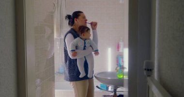 Anne bebeğini banyoda tutarken dişlerini fırçalıyor. Bebek, sabah rutinine ilgi göstererek çevreye merakla bakıyor.