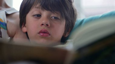 Büyük çocuğun kitap okuduğu yakın plan, annesinin yanında otururken derinden odaklanmış, hikaye anlatırken huzurlu bir aile anı, ev ortamı yaratıyor.