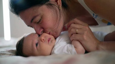 Ebeveyn bebeği yakın tutuyor ve yatakta yanaktan öpüyor, bebek yumuşak bir ifadeyle yukarı bakıyor, duygusal bağlantı, bakım ve anne-baba ile bebek arasındaki derin bağ.