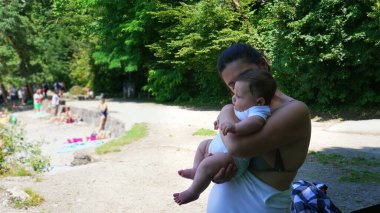 Anne, kumsalda bir göl kıyısında dururken bebeğini kucağında tutuyor. Uzağa bakıyor. İnsanlar su kenarında rahatlarken.