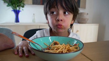 Yanakları dolu bir şekilde spagetti yiyen genç bir çocuk evde rahat bir aile yemeği sırasında sağlıklı bir yemeğin tadını çıkarıyor.