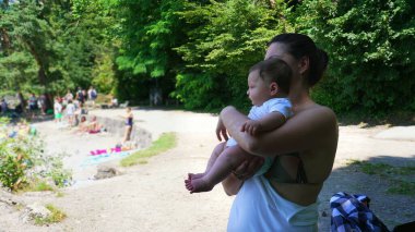 Anne, kumsalda bir göl kıyısında dururken bebeğini kucağında tutuyor. Uzağa bakıyor. İnsanlar su kenarında rahatlarken.