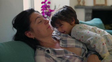Anne kanepeye uzanmış, oğlu üzerinde dinlenirken hafifçe gülümsüyor huzurlu bir evde sevgi dolu bir anı paylaşıyor.