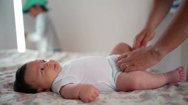 Bebek yatakta yatarken, ebeveyn giysilerini ayarlar, arka planda kardeşlik oynar, huzurlu ve sakin bir aile ortamı kurar, ev ortamında bağ ve günlük anları gösterir.