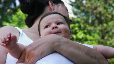Anne bebeğini dışarıda tutarken, bebek kollarında dinlenirken dikkatlice bakarken, yeşillik ve doğa ile çevriliyken.