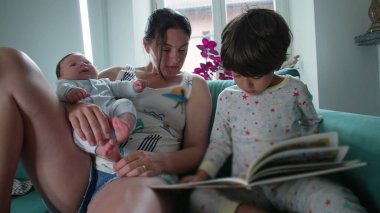 Anne, kucağında bebeği tutarken oğluyla kitap okuyor, kanepede sıcak bir aile anı yaratıyor, beraberlik gösteriyor, öğreniyor ve bağ kuruyor.