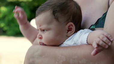 Doğayla ve yeşilliklerle çevrili bir yere dikkatlice bakarken annelerin kollarında dinlenen bir bebeğin profil görüntüsü.