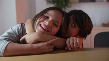 Genç bir kızın, masada oturmuş kardeşine sarılırken, hem gülümseyip hem de şefkat gösterirken, güçlü bir kardeş bağının altını çizerken.