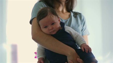 Anne iki eliyle bebeği tutuyor, bebeğin bacaklarını ve ayaklarını rahat bir pozisyonda gösteriyor, ebeveyn ve çocuk arasındaki nazik destek ve ilgiye odaklanıyor.