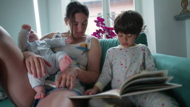 Anne, kucağında bebeği tutarken oğluyla kitap okuyor, kanepede sıcak bir aile anı yaratıyor, beraberlik gösteriyor, öğreniyor ve bağ kuruyor.