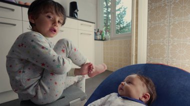 Pijamalı çocuk mutfakta oturuyor ve evde sakin bir anda bebek kardeşiyle kibarca etkileşime geçiyor. Ev ortamında kardeşler arasındaki aile bağı ve bakımı
