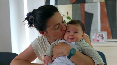 Mutlu anne gülümseyen bebeğini kucağında tutuyor, her ikisi de sıcak bir ev ortamında neşeli bir anı paylaşıyor. Anne-çocuk iç açıcı bir sahnede kaynaşıyor.