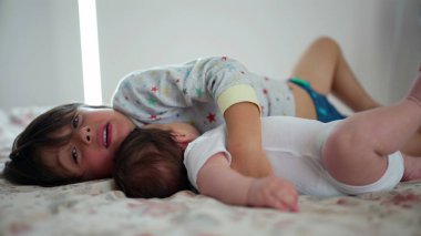 Küçük çocuk yatakta küçük kardeşine sarılıyor, ev ortamında kardeşler arasında tatlı ve neşeli bir an yaratıyor, sevgi, sevgi ve iki çocuk arasında bir bağ olduğunu gösteriyor.