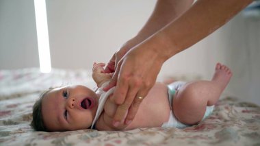 Ebeveyn bebeği yatakta nazikçe giydirirken bebek yukarı bakar, kollar yukarı, doğal duygularla dolu samimi bir ev ortamı, bakım, rahatlık ve bağlanma anlarına odaklanır.