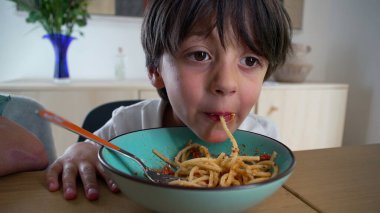 Yanakları dolu bir şekilde spagetti yiyen genç bir çocuk evde rahat bir aile yemeği sırasında sağlıklı bir yemeğin tadını çıkarıyor.