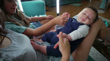 Mother and daughter holding and gently interacting with a baby in their lap, sharing an intimate moment of bonding and connection in a cozy home setting clipart