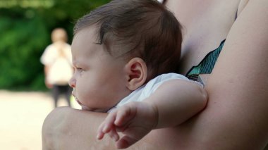 Doğayla ve yeşillikle çevrelenmiş bir şekilde bir şeye dikkatle bakarken annesinin kollarında dinlenen bebeğin profili.