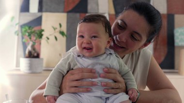 Mutlu bebeğini kapalı bir ortamda kucaklayan gülümseyen anne birlikte neşeli ve şefkatli bir anı paylaşıyor. Anne ve çocuk arasındaki sıcak aile etkileşimi