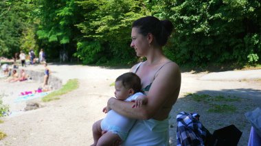 Anne, kumsalda bir göl kıyısında dururken bebeğini kucağında tutuyor. Uzağa bakıyor. İnsanlar su kenarında rahatlarken.