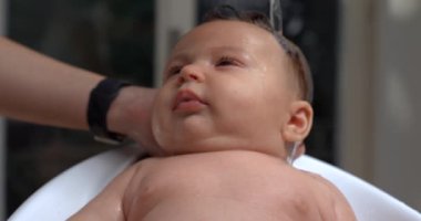 Bebek, banyo sırasında su dökülürken yukarı bakıyor, yüzüne su damlacıkları dökülüyor, sakin ve özenli, banyo bakımı anı. 800 fps yavaş çekim