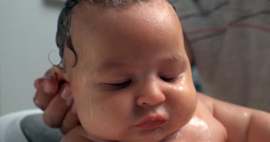 Banyo sırasında duruladıktan sonra bebeğe yakın çekim, bebek yüzünde su, bebek sakin ve rahat, ebeveyn bebek başını nazikçe tutuyor, hassas banyo bakımı anları. 800 fps yavaş çekim