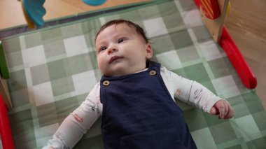 Bebek oyun hasırına uzanmış, merakla yukarı bakıyor. Bebek evde güvenli ve rahat bir ortamda yatarken çevreyi gözlüyor.