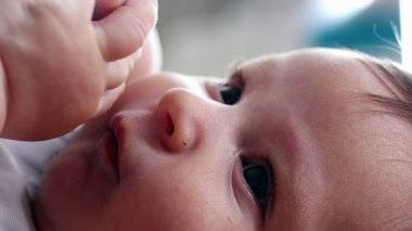 Yatakta dinlenirken kendi elini tutan bir bebeğin yakın çekimi. Yüzün yanındaki bebek eline odaklan. Yumuşak cildi ve bebeğin doğal refleksini vurgula.