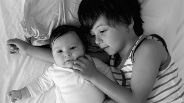 Bebeğin siyah-beyaz yakın çekimi ve yatağında yatan kardeş, sıcak bir aile ortamında hassas ve samimi bir anda bebeğin yüzüne nazikçe dokunuyor.