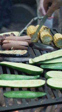 Grilling zucchini slices, corn on the cob, sausages, and skewered meat over an open flame at a campfire. Mixed variety of food grilling on a barbeque in an outdoor forest setting clipart