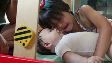 Genç bir çocuk yerde bir bebeğin yanında yatıyor, evde oyun sırasında bebekle nazikçe iletişim kurarken şefkat ve bağlılık gösteriyor.