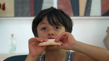 Yemek masasında tapyoka yiyen genç bir çocuk, iki eliyle yemeği tutarken büyük bir ısırık alıyor, evde rahat bir yemeğin tadını çıkarıyor.