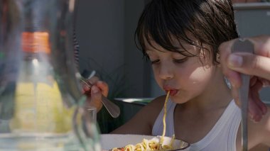 Genç çocuk spagetti yerken uzun bir erişteyi şapırdatarak yiyor, makarnayı çatalla çeviriyor ve gün boyunca dışarıda bir masada yemeğin tadını çıkarıyor.