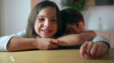 Genç bir kız ve küçük bir oğlan masada otururken birbirlerine sarılıyor, sıcak bir kardeş bağı sergiliyor, her ikisi de gülümsüyor.