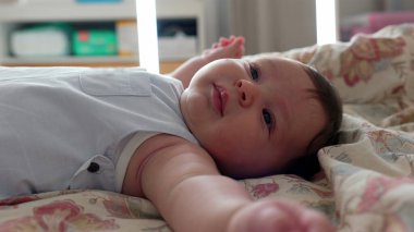 Bebek yatakta gözleri açık yatıyor, kolunu kameraya doğru uzatıyor. Rahat bir evde sakin ve rahat bir an.
