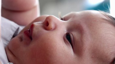 Yatakta dinlenirken kendi elini tutan bir bebeğin yakın çekimi. Yüzün yanındaki bebek eline odaklan. Yumuşak cildi ve bebeğin doğal refleksini vurgula.