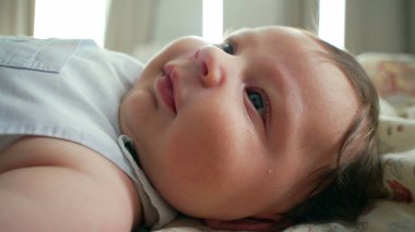 Yatakta uzanmış, yumuşak bir gülümsemesi ve parlak gözleri olan bir bebeğin yakın çekimi. Kapalı mekan sahnesi bebeğin rahat ve neşeli tavrına odaklanıyor.