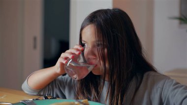 Yemek masasında bardaktan su içen genç bir kız, yemek saatinde bardağı tutarken ve su içerken yüzündeki konsantre ifadeye odaklanıyor.