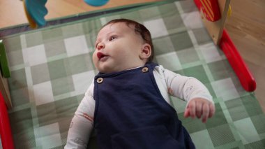 Bebek oyun hasırına uzanmış, merakla yukarı bakıyor. Bebek evde güvenli ve rahat bir ortamda yatarken çevreyi gözlüyor.