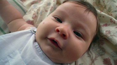 Bebek battaniyeye uzanıp nazikçe kameraya bakıyor. Sahne bebek yüzüne ve vücudunun üst kısmına odaklanıyor.