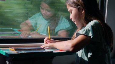 Hareket halindeki bir trenin penceresinde otururken çizimine konsantre olan genç kız, odaklı bir sanat eseri yaratırken penceredeki yansıması görülebilir.