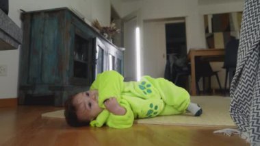 Bebek rahat bir evde yerde yatıyor, yeşil bir kıyafet giyiyor, garip bir şekilde etrafı gözetliyor, paspasın üzerinde huzurlu ve mutlu bir an yaşıyor.