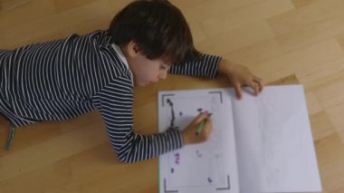 Yerde yatan genç bir çocuk, yeşil kalemle çizim yapmaya odaklanmış, yaratıcı aktivitelere yoğun bir şekilde odaklanmış, çocukluk yaratıcılığı, kapalı alanda oyun zamanı
