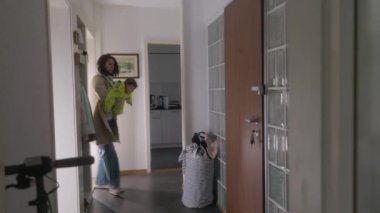 Anne bebeğini kucağında tutarken evden ayrılıyor, sıcak yeşil bir kıyafet giyiyor, dışarı çıkmaya hazırlanıyor, rahatlatıcı ve koruyucu bir jest yapıyor.