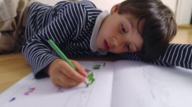 Yeşil kalemle çizim yaparken başını dinlendiren genç bir çocuk, çizim sayfasında yeşil renkleri doldurmaya odaklanmış, yaratıcı aktivitelerde çocukluk ilişkisi