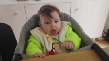 Anne bebeğini beslerken büyük çocuk kendi yemeğine odaklanıyor, aile yemek masasında birlikte yemek yiyor, evde ebeveynlik yapıyor, bebek yüksek sandalyede yiyor, bağ kuruyor.