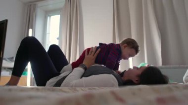 Anne yatakta uzanıyor, bebek üstte dinleniyor, neşeli ve samimi bir aile anı, sıcak bir ev ortamında bağ kuruyor ve rahatlıyor, birlikte vakit geçiriyoruz.