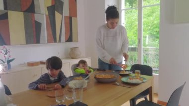 Anne yemek masasında yemek servisi yaparken, büyük çocuk ve bebek bakıcılığı, aile yemeği hazırlama ve sıcak bir evde toplanma.