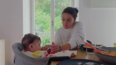 Anne yüksek sandalyede bebeği dikkatlice besliyor, bebek annesinin yardımıyla yemek yiyor, yemek masasında samimi beslenme anı, odaklanmış anne, bakım etkileşimi